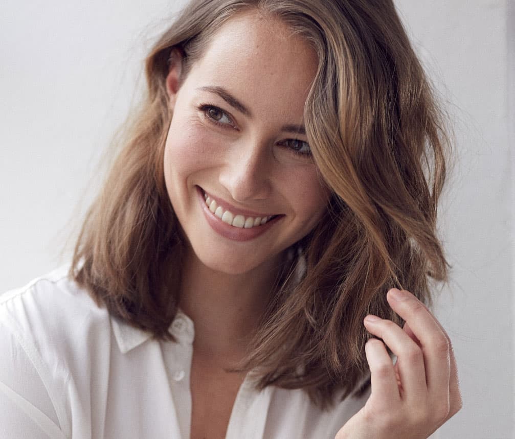 Smiling young woman with long, wavy brown hair wearing a white button-down shirt.