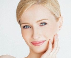 Smiling blonde woman with short hair, fair skin, and blue eyes who appears to be touching her face.